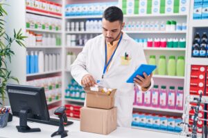 Young hispanic man pharmacist using touchpad holding pills bottle at pharmacy