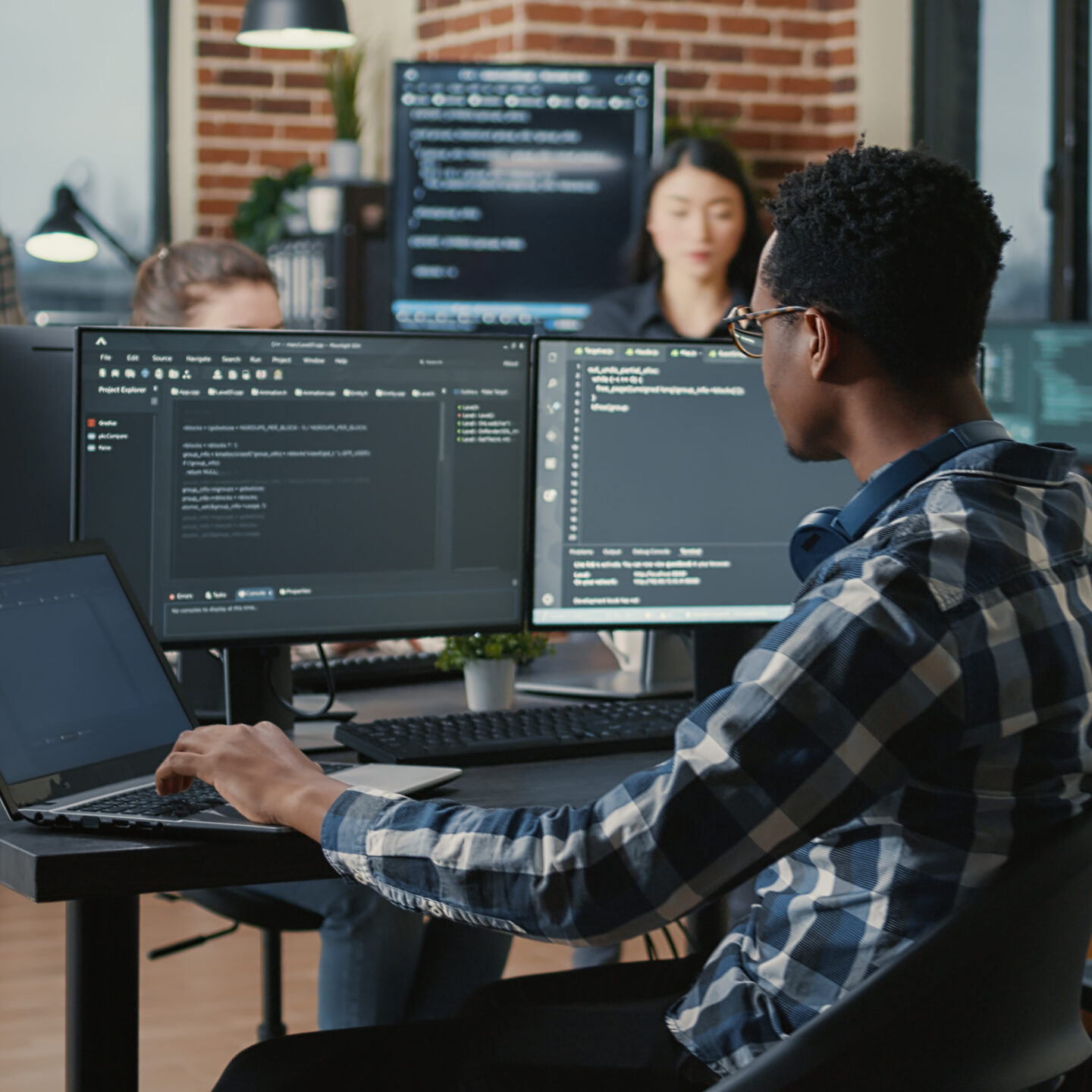 Portrait of system engineer fixing glasses while typing on laptop smiling satisfied about compiling results sitting at desk with multiple screens. App developer enjoying working on user interface.